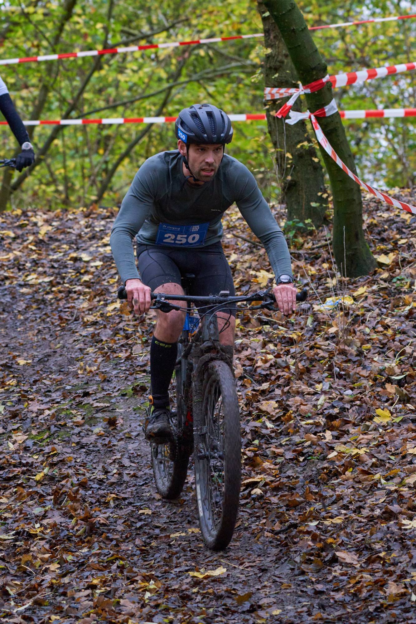 Edwin riding a muddy mountain bike down a leaf-covered forest trail during a race, wearing bib number 250.